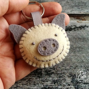 May include: A handmade felt pig keychain. The pig has a light beige face with gray ears and snout. The details are stitched with gray thread, and it has a small gray ribbon loop attached to a silver keyring. The keychain is a cute accessory.