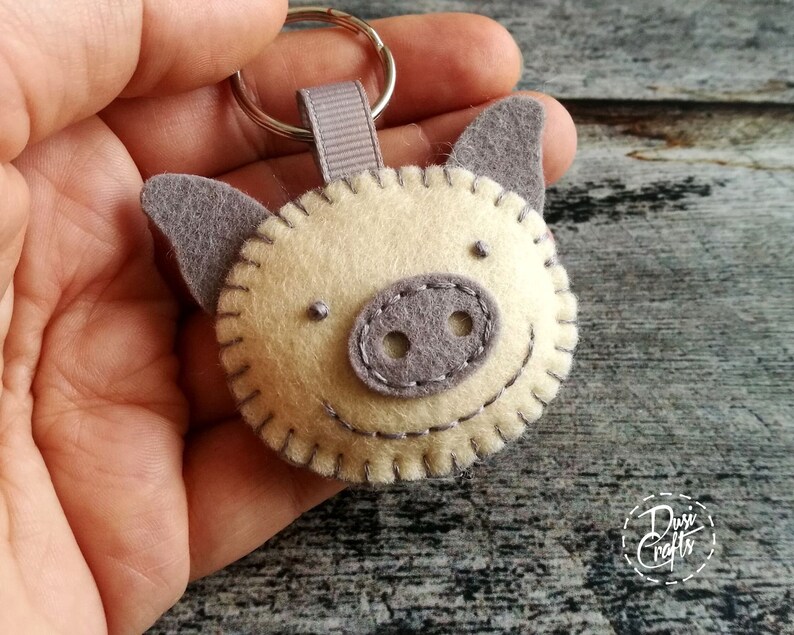 May include: A handmade felt pig keychain. The pig has a light beige face with gray ears and snout. The details are stitched with gray thread, and it has a small gray ribbon loop attached to a silver keyring. The keychain is a cute accessory.
