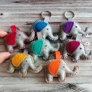 May include: A collection of six felt elephant keychains, each featuring a grey body with a distinctively colored ear in red, blue, purple, teal, magenta, or orange. Each elephant is attached to a metal keyring by a small fabric ribbon loop.