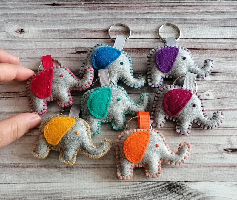 May include: A collection of six felt elephant keychains, each featuring a grey body with a distinctively colored ear in red, blue, purple, teal, magenta, or orange. Each elephant is attached to a metal keyring by a small fabric ribbon loop.