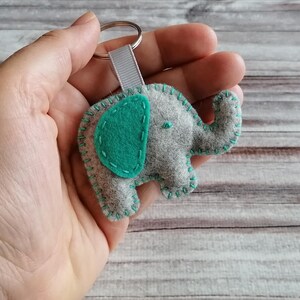 May include: A gray felt elephant keychain featuring a teal felt ear and teal blanket stitching along the edges. The elephant is attached to a silver metal split ring by a short, light gray ribbed fabric ribbon loop for secure key attachment.