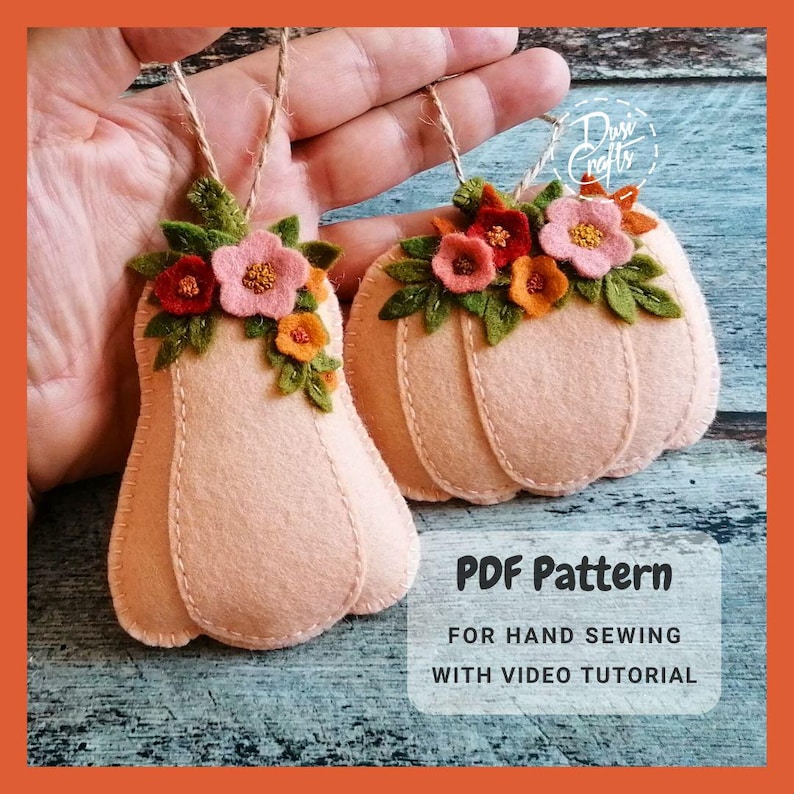 May include: Two felt pumpkin ornaments with floral embellishments. The pumpkins are peach colored and have a stitched stem. The flowers are pink, orange, and green. The ornaments are hanging from twine. The text "PDF Pattern" is below the ornaments. The text "For Hand Sewing" is below the "PDF Pattern" text. The text "With Video Tutorial" is below the "For Hand Sewing" text.