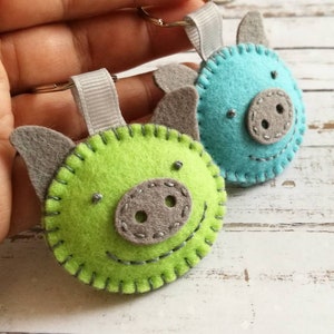 May include: Two felt pig keychains. One is lime green, the other is turquoise. Both have gray ears and snouts, with stitched details for eyes and smiles. Each keychain has a gray ribbon loop and a silver keyring.