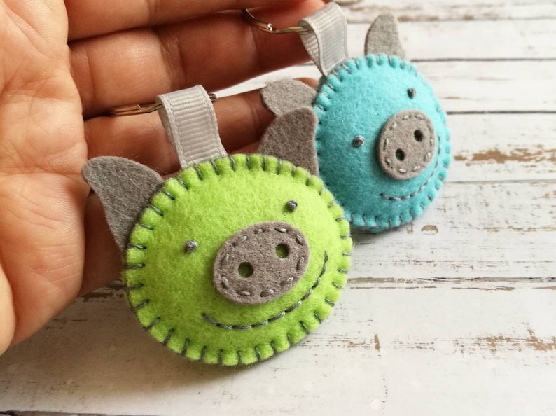 May include: Two felt pig keychains. One is lime green, the other is turquoise. Both have gray ears and snouts, with stitched details for eyes and smiles. Each keychain has a gray ribbon loop and a silver keyring.