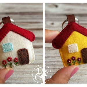 May include: A felt house keychain with a red roof, brown door, and embroidered flowers. The house has a light blue window and a brown ribbon loop for the keyring. The second house is yellow with a red roof.