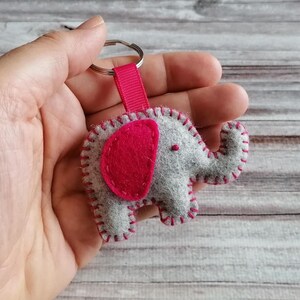 May include: A gray felt elephant keychain featuring a bright pink felt ear and matching pink stitching along the edges. The elephant is attached to a silver metal split ring by a short, ribbed pink fabric ribbon loop.