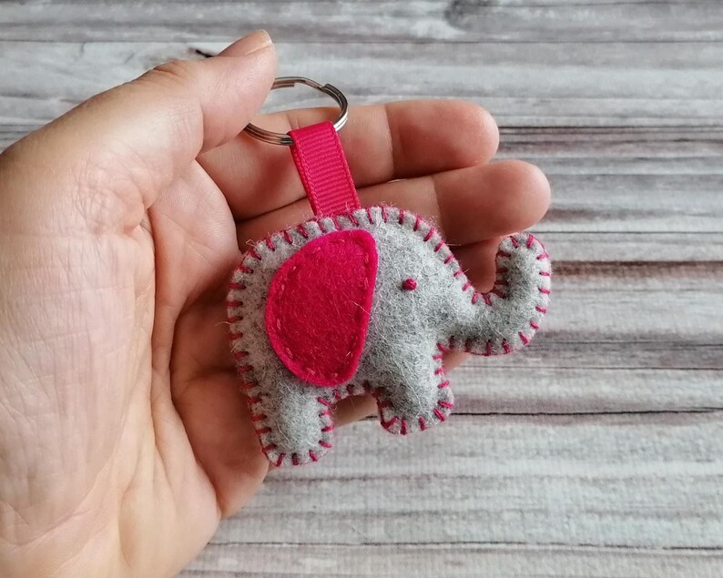 May include: A gray felt elephant keychain featuring a bright pink felt ear and matching pink stitching along the edges. The elephant is attached to a silver metal split ring by a short, ribbed pink fabric ribbon loop.