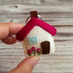 May include: A handmade felt house keychain with a white body, a red roof, and a brown door. The house has a light blue window and embroidered pink flowers. A brown ribbon loop is attached to a silver keyring.