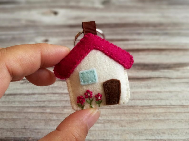 May include: A handmade felt house keychain with a white body, a red roof, and a brown door. The house has a light blue window and embroidered pink flowers. A brown ribbon loop is attached to a silver keyring.