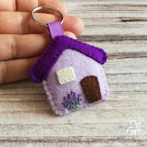 May include: A handmade felt house keychain in shades of purple and lavender. The house features a dark purple roof, a brown door, and a white window. A small embroidered lavender bush is below the door. The keychain is attached to a silver ring.