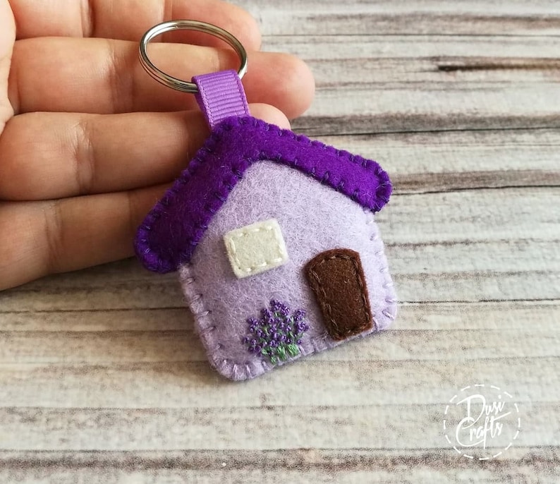 May include: A handmade felt house keychain in shades of purple and lavender. The house features a dark purple roof, a brown door, and a white window. A small embroidered lavender bush is below the door. The keychain is attached to a silver ring.