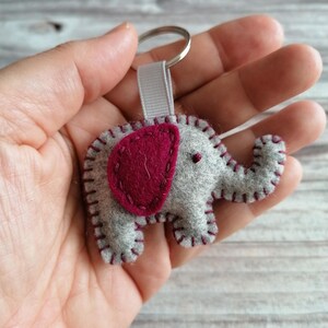 May include: A gray felt elephant keychain featuring a dark pink felt ear, dark pink blanket stitching along the edges, and a small dark pink embroidered eye. The elephant is attached to a silver metal split ring by a light gray fabric ribbon loop.