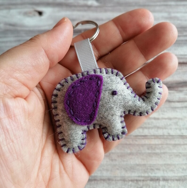 May include: A gray felt elephant keychain featuring a dark purple felt ear and dark purple blanket stitching along the edges. The elephant is attached to a silver metal split ring by a small gray ribbon loop, creating a compact animal accessory.