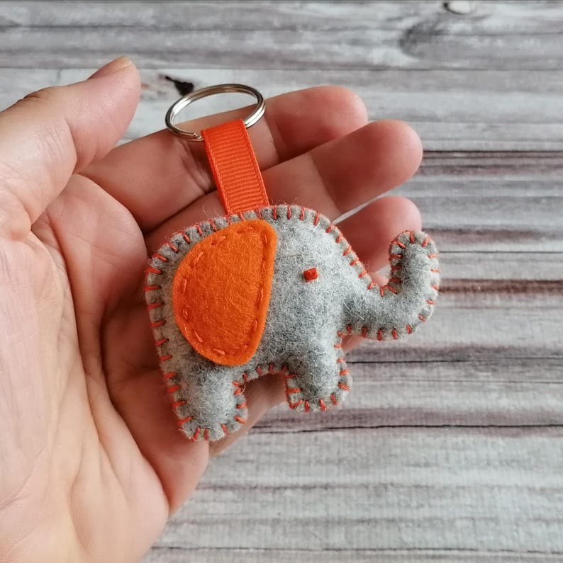 May include: A gray felt elephant keychain featuring a bright orange felt ear and matching orange blanket stitching along the edges. The elephant is attached to a silver metal split ring by a short orange fabric ribbon loop.