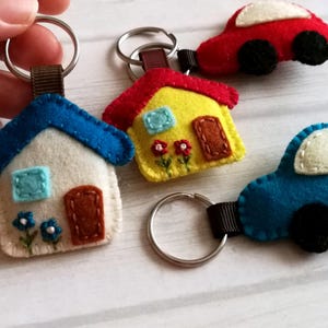 May include: A collection of handmade felt keychains in the shape of a house and cars. The house keychains are white with blue roofs, and the cars are red and blue. Each keychain has a metal ring and a brown fabric loop.