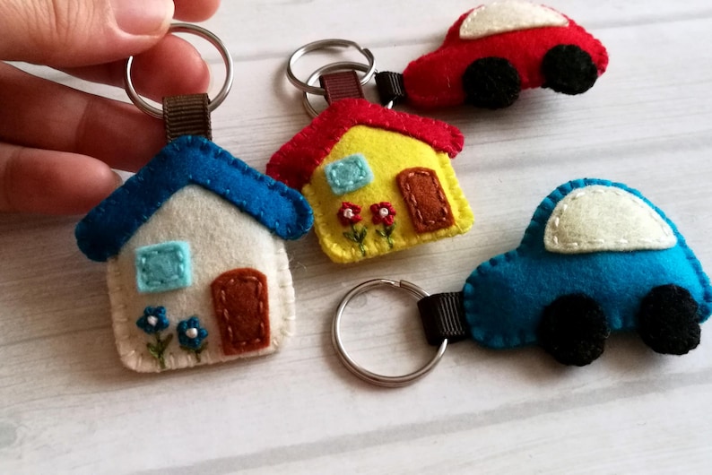 May include: A collection of handmade felt keychains in the shape of a house and cars. The house keychains are white with blue roofs, and the cars are red and blue. Each keychain has a metal ring and a brown fabric loop.