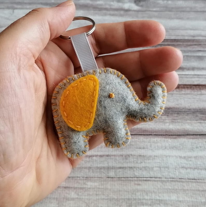 May include: A gray felt elephant keychain featuring a bright yellow felt ear, yellow blanket stitching along the edges, and a small yellow eye. The elephant is attached to a silver metal split ring by a short, light gray fabric ribbon loop.