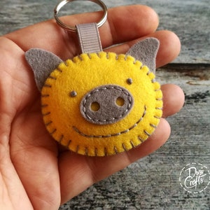 May include: A handmade yellow felt pig keychain with gray ears and snout. The pig has a stitched smile and is attached to a silver keyring with a gray ribbon loop. The keychain is a cheerful accessory.