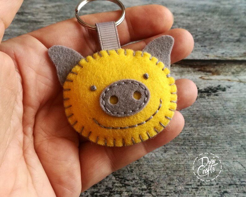 May include: A handmade yellow felt pig keychain with gray ears and snout. The pig has a stitched smile and is attached to a silver keyring with a gray ribbon loop. The keychain is a cheerful accessory.