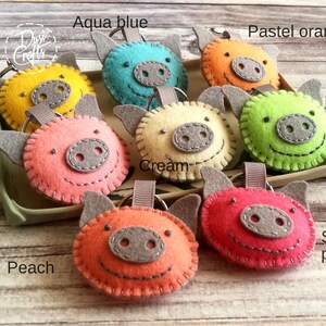 May include: A collection of handmade felt pig keychains in various colors, including yellow, aqua blue, pastel orange, green, soft pink, cream, peach, and strawberry pink. Each keychain features a gray snout, ears, and a stitched outline.
