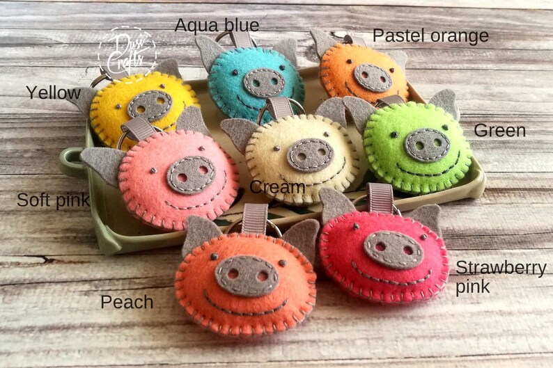May include: A collection of handmade felt pig keychains in various colors, including yellow, aqua blue, pastel orange, green, soft pink, cream, peach, and strawberry pink. Each keychain features a gray snout, ears, and a stitched outline.