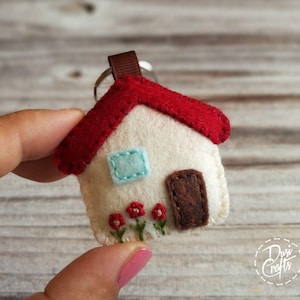 May include: A handmade felt house keychain with a red roof, cream-colored walls, and a brown door. The house features a light blue window and embroidered red flowers. A brown ribbon and metal ring are attached.