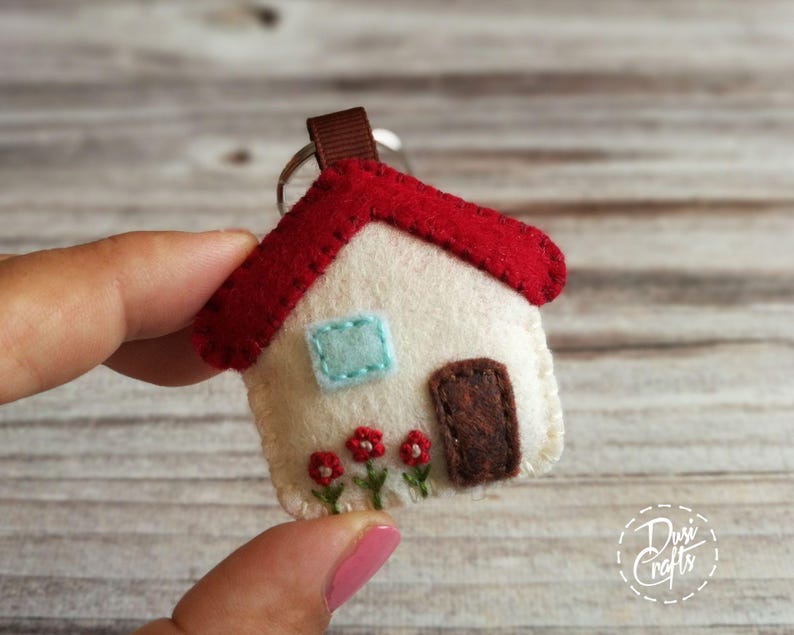 May include: A handmade felt house keychain with a red roof, cream-colored walls, and a brown door. The house features a light blue window and embroidered red flowers. A brown ribbon and metal ring are attached.