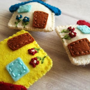 May include: Close-up of three handmade felt house ornaments. Each house features a different color scheme, including yellow, white, and red, with embroidered details like flowers and doors. The ornaments are designed with a whimsical, handcrafted aesthetic.
