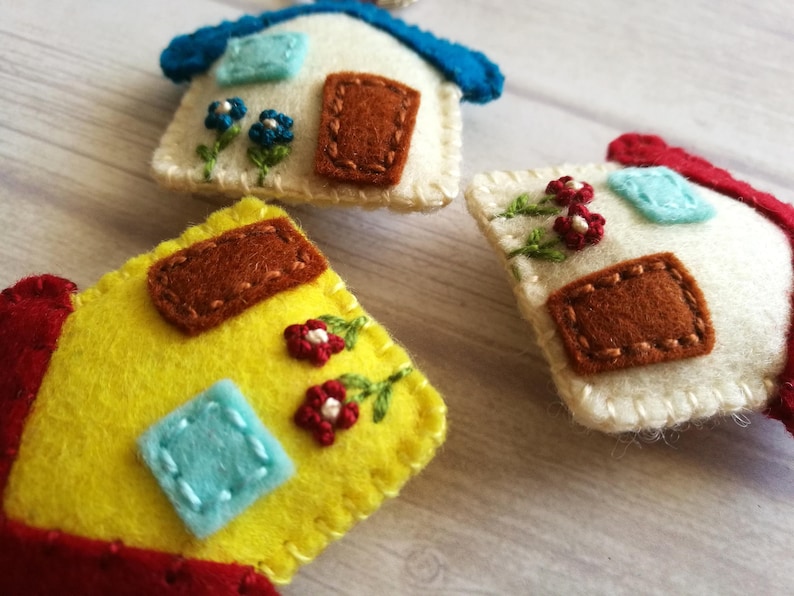 May include: Close-up of three handmade felt house ornaments. Each house features a different color scheme, including yellow, white, and red, with embroidered details like flowers and doors. The ornaments are designed with a whimsical, handcrafted aesthetic.