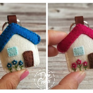 May include: A felt house keychain with a blue roof, white walls, and a brown door. The house has a small blue window and embroidered flowers. A similar keychain has a pink roof. Both are held up by a brown ribbon and a silver keyring.