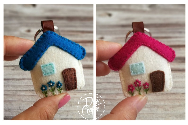 May include: A felt house keychain with a blue roof, white walls, and a brown door. The house has a small blue window and embroidered flowers. A similar keychain has a pink roof. Both are held up by a brown ribbon and a silver keyring.