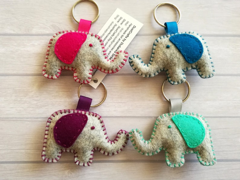 May include: Four felt elephant keychains in gray with colorful ears and matching stitching. The colors include hot pink, blue, dark purple, and teal. Each elephant has a metal keyring attached by a ribbon loop. A white tag with black text is attached.