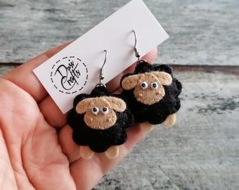 以下が含まれることがあります： 黒い羊毛の体とベージュの顔、白と黒の目を持つ、手作りのフェルト製羊のイヤリング。羊は銀色の金属製イヤリングフックに取り付けられており、Dusi Craftsという文字が印刷された白い台紙に固定されています。