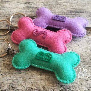 May include: Three felt dog bone keyrings in pink, purple, and teal. Each bone has a stitched letter and a metal keyring. The keyrings are handmade and feature a decorative stitch around the edges.