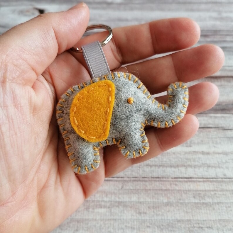 May include: A gray felt elephant keychain featuring a bright yellow ear and decorative yellow stitching along the edges. The elephant is attached to a silver metal split ring by a small gray fabric loop, creating a compact and tactile accessory.