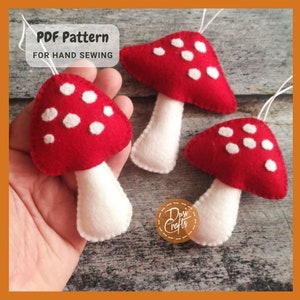 Può includere: Tre decorazioni a forma di fungo in feltro con pois bianchi su sfondo rosso e steli bianchi. Le decorazioni sono appese a una corda. Il testo "PDF Pattern for Hand Sewing" è visibile nell'immagine.
