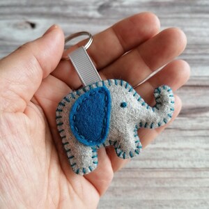 May include: A gray felt elephant keychain featuring a dark blue felt ear, decorative teal stitching along the edges, and a light gray ribbon loop attached to a silver metal split ring. The elephant has a small dark blue dot for an eye.