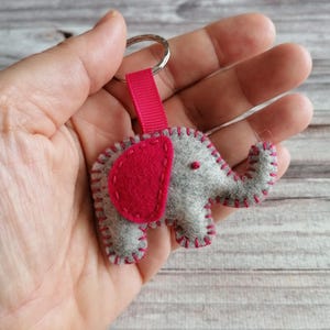 May include: A gray felt elephant keychain featuring a bright pink felt ear and matching pink blanket stitching along the edges. The elephant is attached to a silver metal split ring by a short, vertical pink fabric ribbon loop.