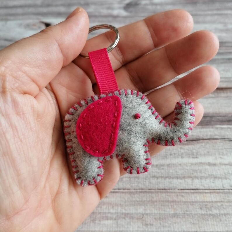 May include: A gray felt elephant keychain featuring a bright pink felt ear and matching pink blanket stitching along the edges. The elephant is attached to a silver metal split ring by a short, vertical pink fabric ribbon loop.