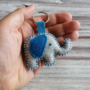 May include: A felt elephant keychain featuring a light gray body with a dark blue ear and a blue fabric loop attached to a silver metal ring. The elephant is constructed with visible blue blanket stitching along the edges and a small blue dot eye.