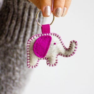 May include: A felt elephant keychain featuring a light gray body with a vibrant magenta ear and matching magenta blanket stitching along the edges. The elephant is attached to a silver metal split ring by a small magenta fabric loop.