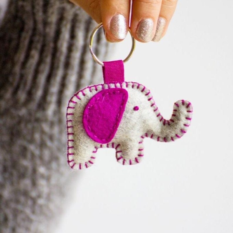May include: A felt elephant keychain featuring a light gray body with a vibrant magenta ear and matching magenta blanket stitching along the edges. The elephant is attached to a silver metal split ring by a small magenta fabric loop.