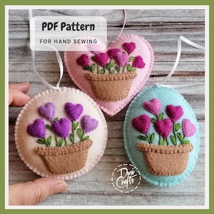 May include: PDF pattern for hand sewing three felt ornaments shaped like a heart, circle, and oval. Each ornament features a brown felt pot with purple felt hearts and green felt stems. The text "PDF Pattern for Hand Sewing" is at the top of the image.