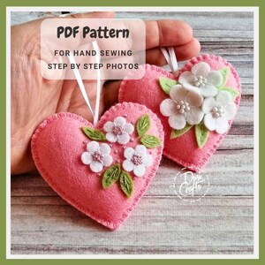 May include: Two pink felt heart ornaments with white flower and green leaf embellishments. The hearts have white ribbon hangers. Text on the image reads "PDF Pattern FOR HAND SEWING STEP BY STEP PHOTOS."
