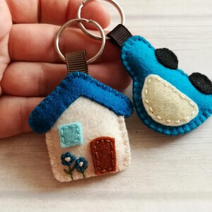 May include: Two handmade felt keychains. One is a white house with a blue roof, a brown door, and blue flowers. The other is a blue bird with black accents. Both have silver key rings and brown ribbon loops.