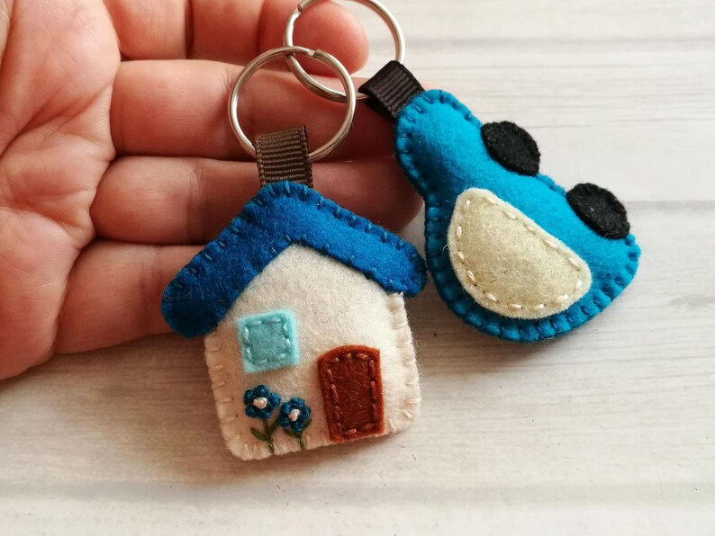 May include: Two handmade felt keychains. One is a white house with a blue roof, a brown door, and blue flowers. The other is a blue bird with black accents. Both have silver key rings and brown ribbon loops.