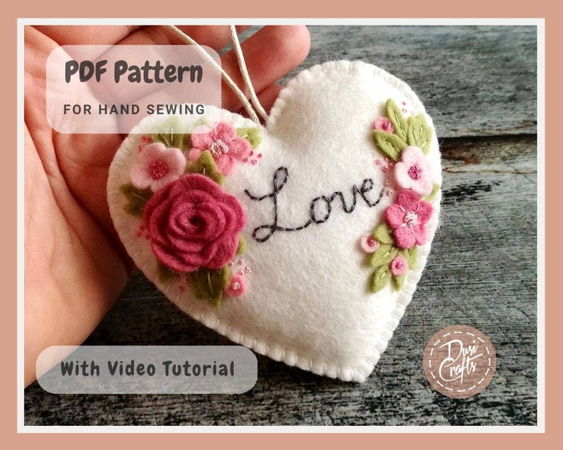 May include: A white felt heart with the word "Love" embroidered in black thread. The heart is decorated with pink and green felt flowers and beads. The image includes the text "PDF Pattern for Hand Sewing" and "With Video Tutorial".