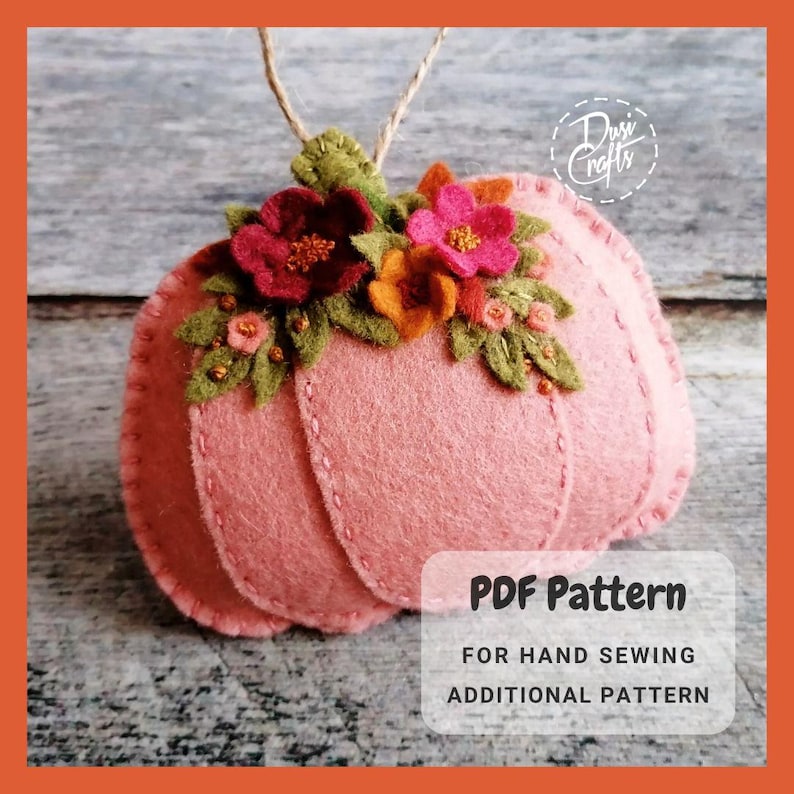 May include: A pink felt pumpkin ornament with a floral design. The pumpkin has a stem and is decorated with felt flowers in shades of red, pink, orange, and green. The ornament is a PDF pattern for hand sewing.