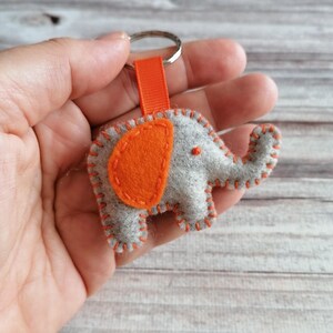 May include: A gray felt elephant keychain featuring a bright orange ear and matching orange blanket stitching along the edges. The elephant is attached to a silver metal split ring by a short, vertical orange fabric ribbon loop.