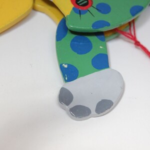 Puede incluir: Un t&iacute;tere de madera de un gato con pintura verde, azul y amarilla. El gato tiene una cuerda roja atada a su espalda.